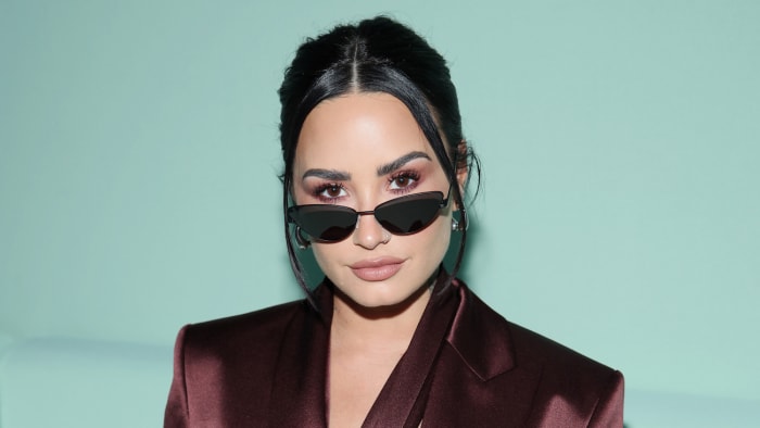 Demi Lovato poses in front of a teal background in a brown satin blazer and sunglasses.